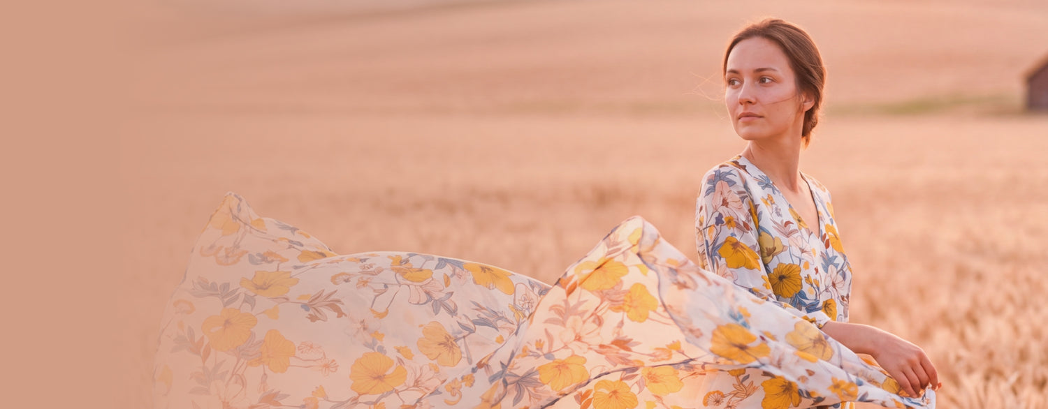 Kobieta w polu zboża trzymająca zwiewną tkaninę w kwiaty — baner główny Textil World, lato 2026
